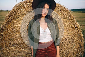 Stylish girl posing at hay bale in summer field in sunset. Portrait of young sensual woman in hat at haystack, atmospheric