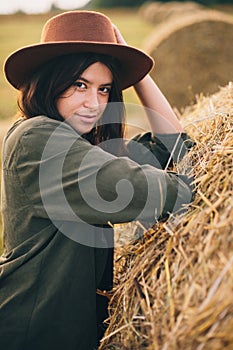 Stylish girl posing at hay bale in summer field in sunset. Portrait of young sensual woman in hat at haystack, atmospheric