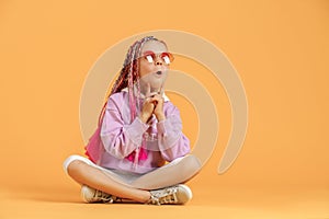 Stylish girl in rounded glasses with pink dreadlocks sitting, thinking, look up and posing on a yellow background