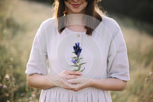 Stylish girl in rustic dress holding wildflower in hand, standing in sunny meadow in mountains. Boho woman gathering gentiana