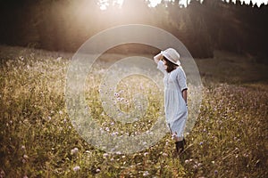 Stylish girl in rustic dress walking in wildflowers in sunny meadow in mountains. Boho woman relaxing in countryside flowers at