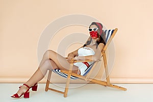 Stylish girl in sunglasses, mask and bathing suit sitting on deck chair