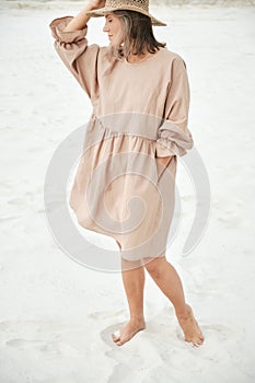 Stylish girl in trendy summer linen dress straw hat posing on the sand. Boho style.