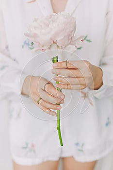 Stylish girl in white floral shirt gently holding peony flower in hands with jewelry. Sensual image. Beautiful young woman holding