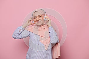 Stylish glamour Arab Muslim woman, wearing a pink hijab, listening to music on wireless headphones and smiling at camera