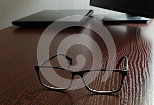Stylish glasses on wooden office table on blurred background of laptop and display
