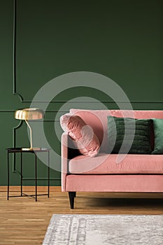 Stylish golden lamp on industrial table next to pink couch with emerald pillows in dark green living room interior,copy space on