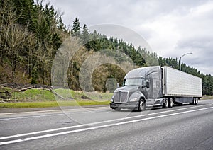 Stylish gray big rig semi truck tractor transporting cargo in refrigerator semi trailer running on the interstate highway road