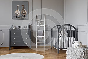 Stylish grey baby room interior with wooden furniture, white scandinavian ladder and teddy bear on pouf, real photo with copy