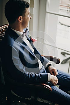 Stylish groom in blue suit, with bow tie and boutonniere with pink rose sitting in chair near window in hotel room. Morning