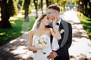 Groom with glasses and bride with a bouquet of peonies gently hug in the park