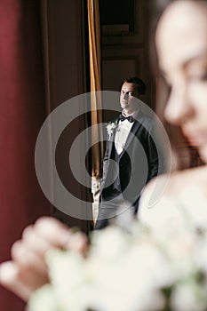 Stylish groom looking at gorgeous bride in amazing dress with bouquet posing at window in luxury room in hotel. rich wedding