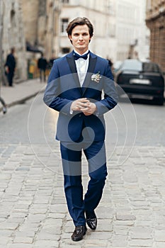 Stylish groom posing on background of old wall in city street. luxury handsome man in blue suit with flowers walking in street