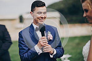 Stylish groom pronouncing vow to his beautiful bride during matrimony. Groom pronouncing speech and holding microphone. Beautiful