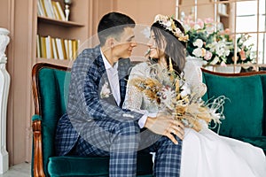 Stylish groom in a suit and a young bride in a wedding lace dress with a bouquet of flowers, sit together, hug, in the studio on