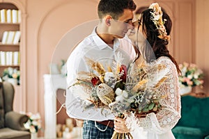 Stylish groom in a suit and a young bride in a wedding lace dress with a bouquet of flowers, sit together, hug, in the studio on