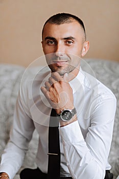 A stylish groom with a watch on his hand and a tie is sitting on a chair in an expensive hotel room.