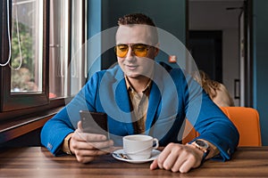 A stylish guy businessman of European appearance in sunglasses, a jacket and shirt sits at a table in a cafe on a coffee break,