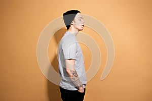 Stylish guy dressed in a gray shirt, black jeans and black hat is posing on the beige background in the studio