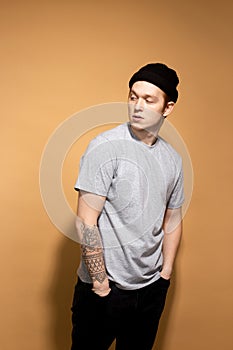 Stylish guy dressed in a gray shirt, black jeans and black hat with tattoo on his hand is posing on the beige background