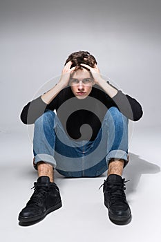 Stylish guy posing in studio sitting on the floor in denim pants and black sweatshirt. Male student portrait full on white