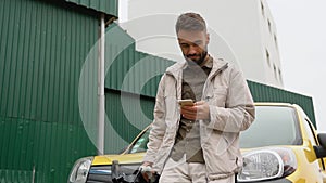 Stylish handsome male standing near modern electric car and texting on smartphone outddors. Young man in good mood