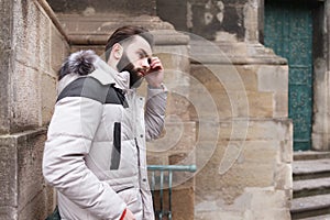 Stylish handsome man with a beard in a winter jacket with fur posing outdoors. Portrait of an elegant guy in beautiful clothes