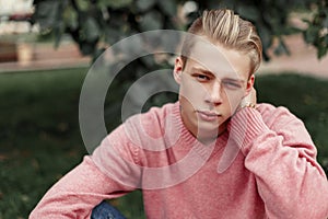 Stylish handsome young man in pink sweater sits in the park.