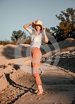 Stylish happy attractive smiling woman posing in desert sand dressed in white clothes wearing straw hat. Travel safari