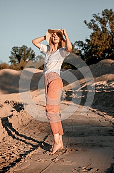 Stylish happy attractive smiling woman posing in desert sand dressed in white clothes wearing straw hat. Travel safari