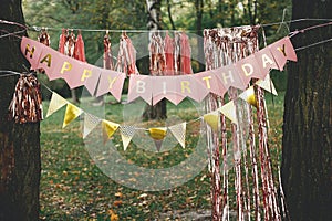 Stylish happy birthday garland hanging in park. Modern rose gold decor, tassel garland and happy birthday banner hanging on trees