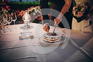 Stylish happy bride and groom cutting together wedding cake with fruits at wedding reception outdoors in the evening. Couple hands