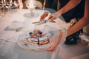 Stylish happy bride and groom cutting together wedding cake with fruits at wedding reception outdoors in the evening. Couple hands