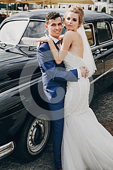 Stylish happy bride and groom embracing at old black retro car. Gorgeous wedding couple of newlyweds hugging after wedding