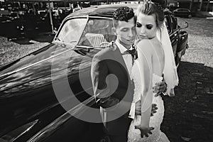 Stylish happy bride and groom embracing at old black retro car. Gorgeous wedding couple of newlyweds hugging after wedding
