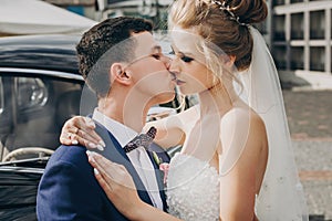 Stylish happy bride and groom kissing at old black retro car. Gorgeous wedding couple of newlyweds embracing after wedding