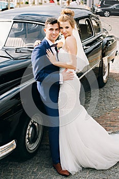 Stylish happy bride and groom posing at old retro car. Gorgeous wedding couple of newlyweds smiling and hugging after wedding