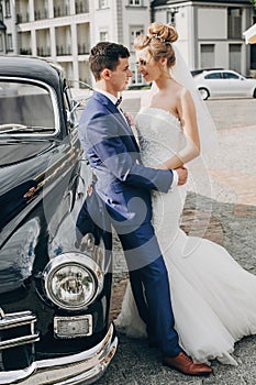 Stylish happy bride and groom posing at old retro car. Gorgeous wedding couple of newlyweds smiling and hugging after wedding