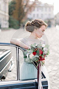 Stylish happy bride standing near the black retro car and holding wedding bouquet. Old city buildings on the backgroud