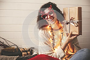 Stylish happy girl in festive glasses with reindeer antlers shaking christmas gift and smiling in christmas lights. Young hipster