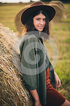Stylish happy girl posing at hay bale in summer field in sunset. Portrait of young sensual woman in hat smiling at haystack,