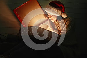 Stylish happy girl in santa hat and cozy sweater opening christmas gift box with magic light on background of presents in dark