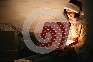 Stylish happy girl in santa hat and cozy sweater opening christmas gift box with magic light on background of presents in dark