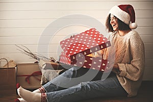 Stylish happy girl in santa hat and cozy sweater opening christmas gift box with magic light on background of presents in modern