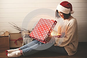 Stylish happy girl in santa hat and cozy sweater opening christmas gift box with magic light on background of presents in modern