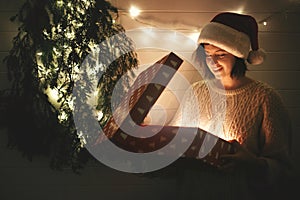 Stylish happy girl in santa hat and cozy sweater opening christmas gift box with magic light in dark room at modern christmas tree