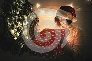 Stylish happy girl in santa hat and cozy sweater opening christmas gift box with magic light in dark room at modern christmas tree
