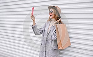 Stylish happy laughing woman taking selfie picture by smartphone with shopping bag in gray coat, round hat posing on city