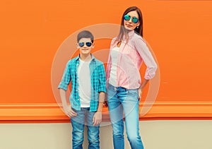 Stylish happy smiling mother with son teenager posing together in sunglasses, checkered shirts, jeans in the city on vivid orange