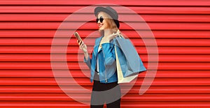 Stylish happy smiling woman with shopping bags and smartphone wearing blue jacket, black round hat on red background